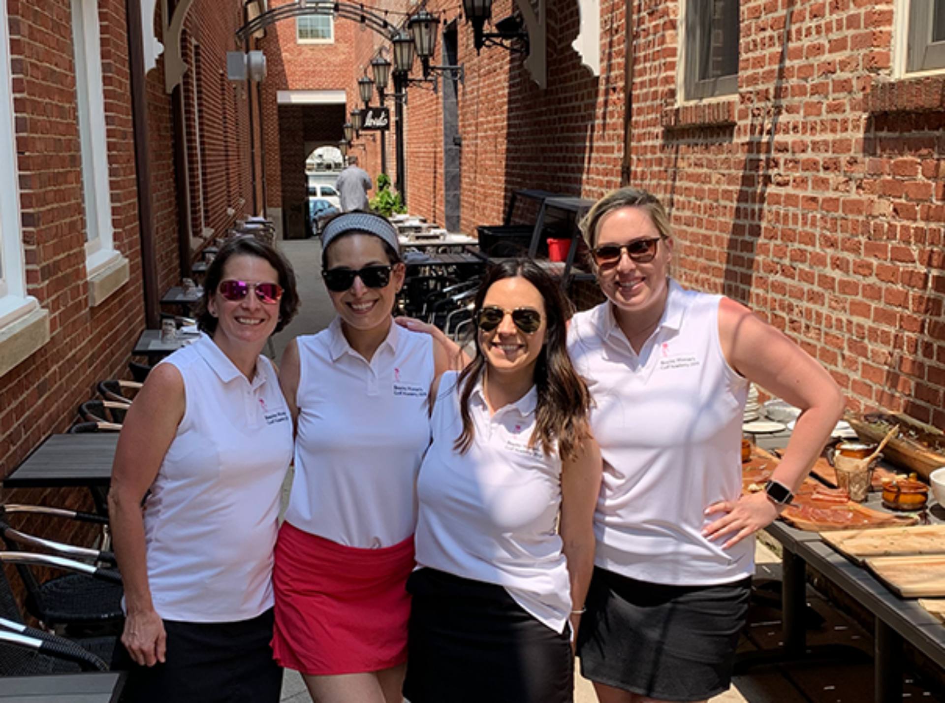 Four women dressed in white shirts and black skirts pose together for a group photo, smiling and looking confident.